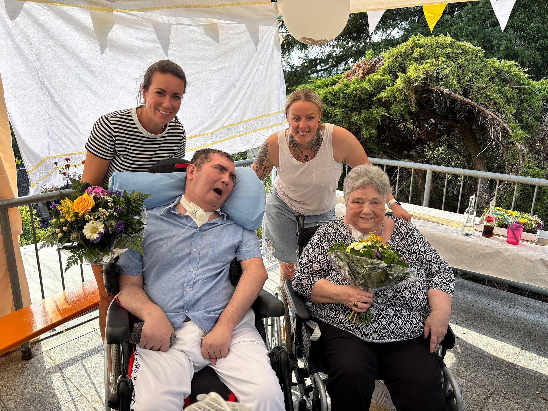 Zwei Pflegekräfte lächeln und überreichen Blumen an Bewohner einer Intensivpflege Wohngemeinschaft im Freien.