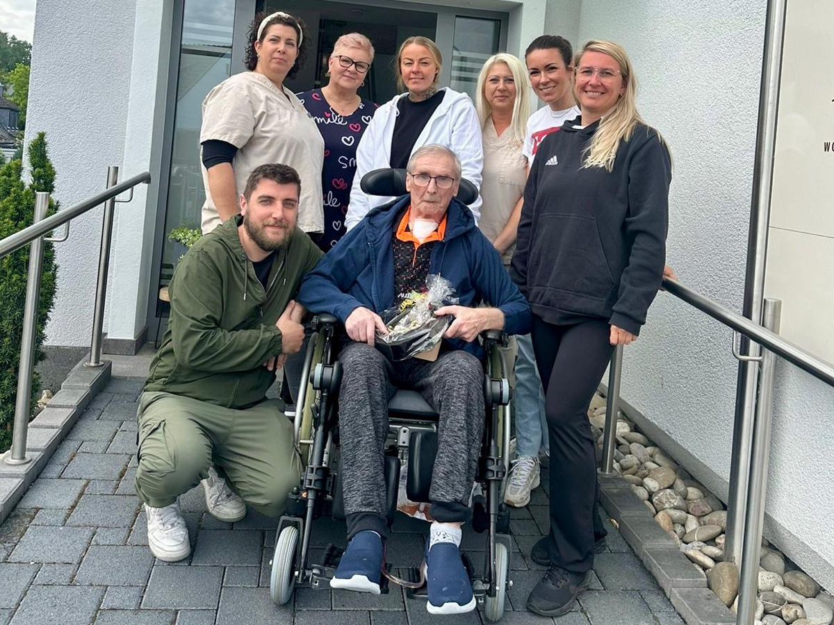 Eine Gruppe von Pflegekräften posiert lächelnd mit einem älteren Mann im Rollstuhl vor einer Außerklinischen Wohngemeinschaft.